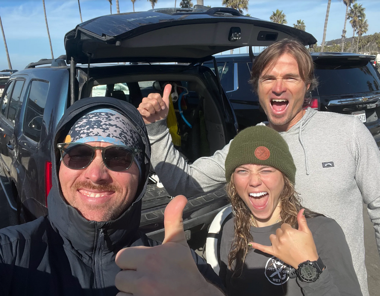 Three smiling divers giving thumbs up and a shaka beside an SUV at the beach