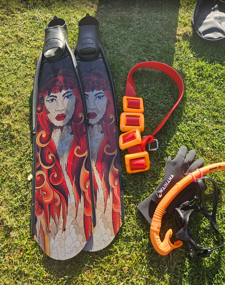Freediving fins with a red-haired mermaid design laid on the grass beside a weight belt, gloves, mask, and orange snorkel