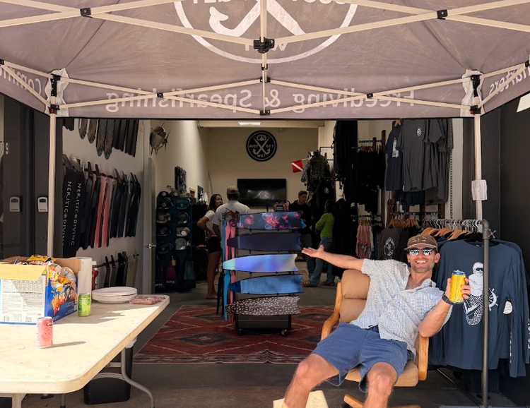 Smiling diver relaxing in a chair under the Just Get Wet tent in front of the shop’s fins and apparel