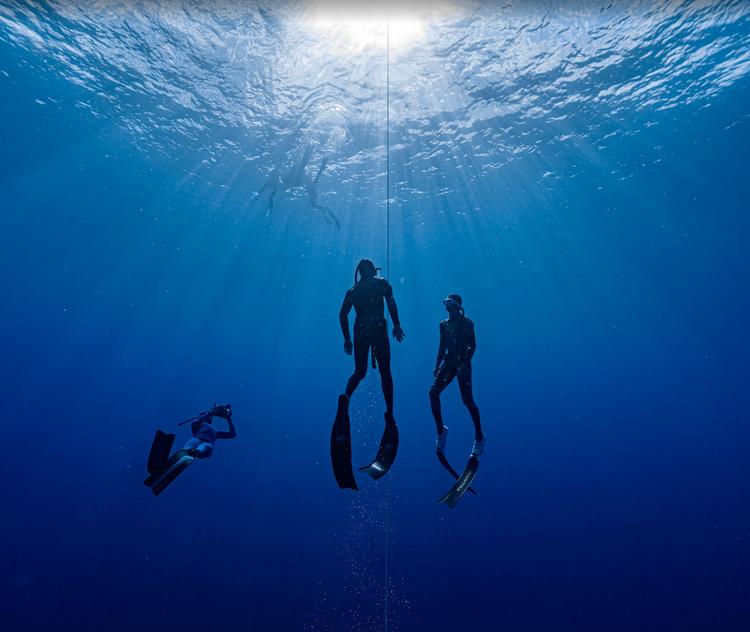 Two freedivers ascending beside a dive line in deep blue water with sunlight streaming from the surface