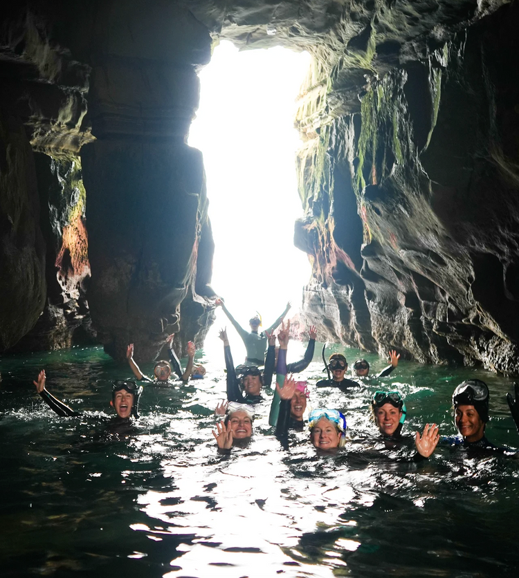 Group of smiling snorkelers waving in the water inside a sea cave lit from the entrance