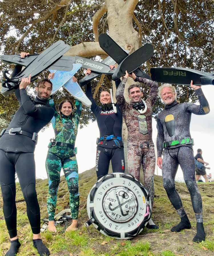 Five smiling freedivers in wetsuits holding their fins overhead beside a Just Get Wet dive buoy
