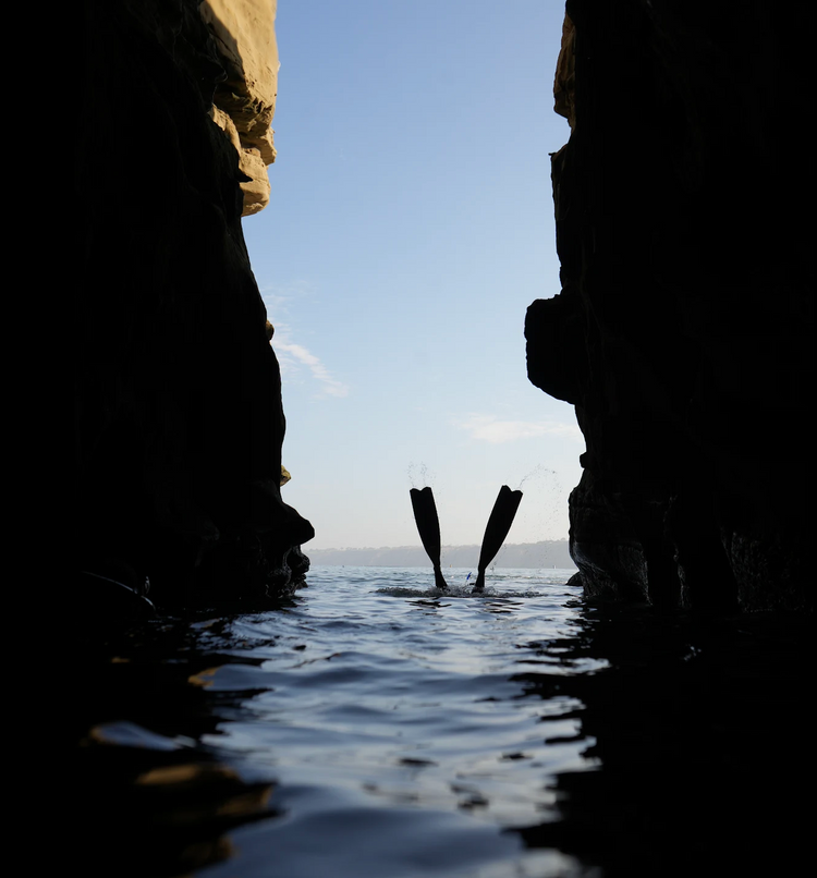 Freediver duck-diving with fins in the air, seen through a narrow sea cave opening